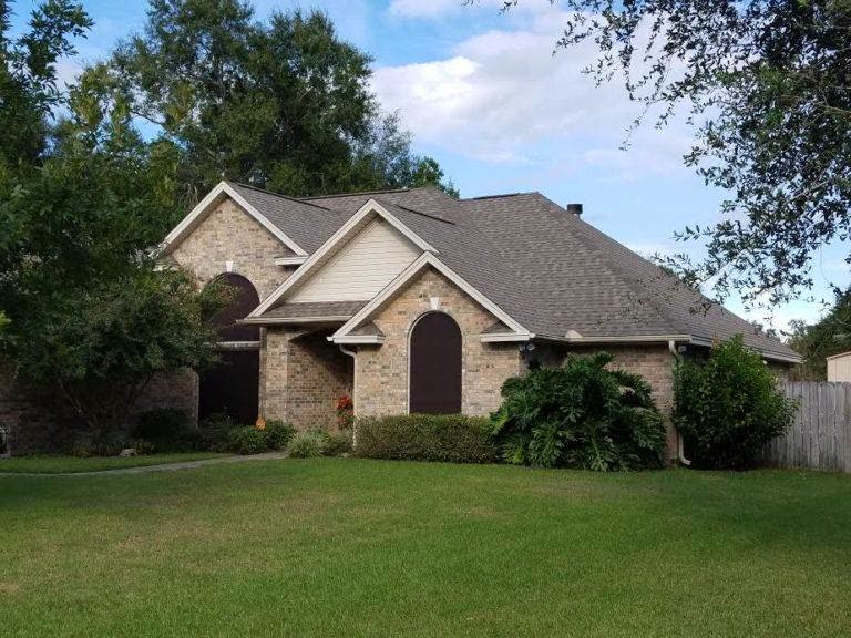 Brick one-story house with stone front and a shingle roof under trees